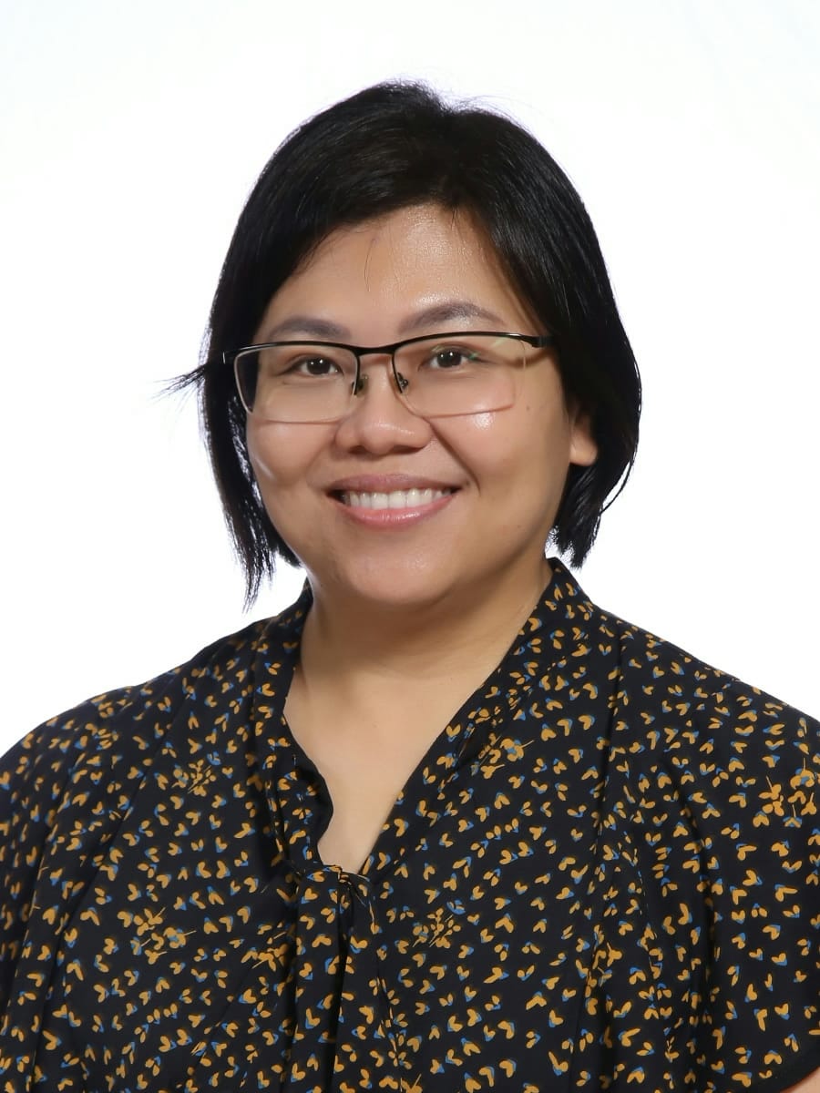 A smiling woman with short black hair and glasses wearing a dark floral patterned blouse against a white background.